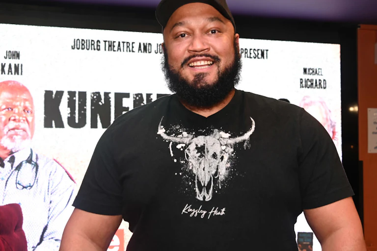 A man with a beard in a cap and T-shirt smiles as he looks at the camera, a theatre banner behind him.
