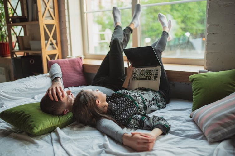 A man and woman lying on a bed watching something on a laptop.