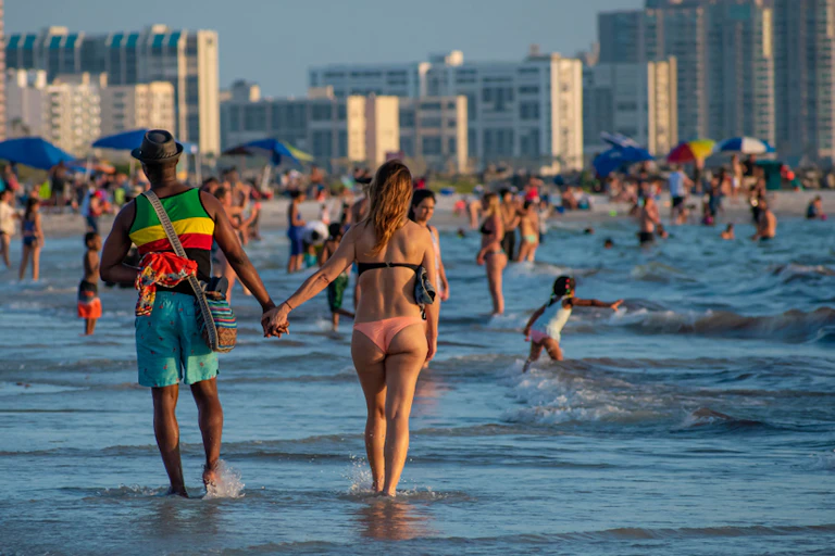 A couple holding hands walk through sea water with a Florida skyline in the background.
