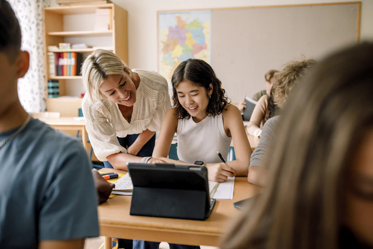 Smiling teacher leans over shoulder of student seated in front of a tablet in a classroom