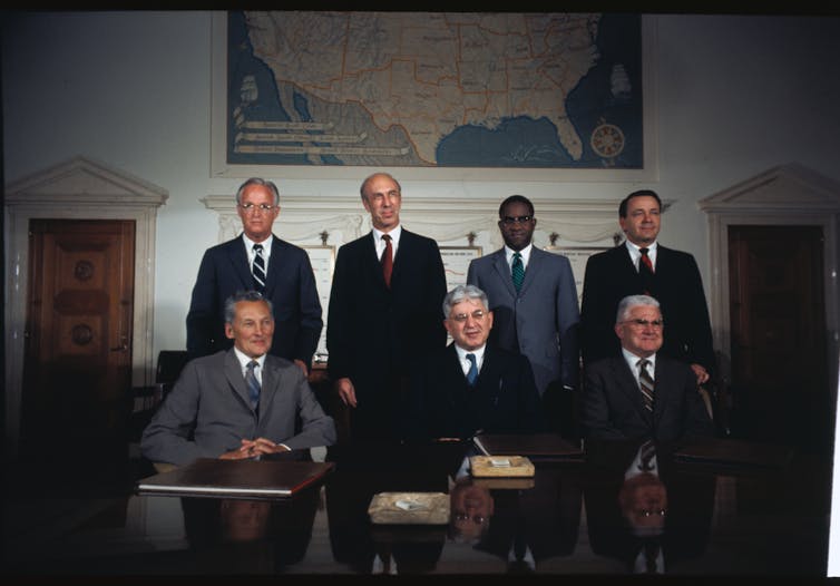 Three men in suits sit side by side at a conference table while four men, also wearing suits, stand behind them. A large map of the US hangs on a wall in the background.