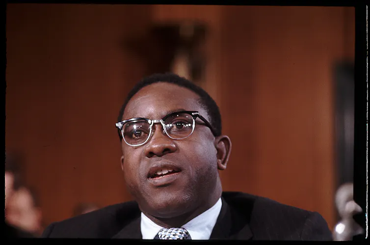 A seated man wearing a suit and glasses is photographed as he speaks.