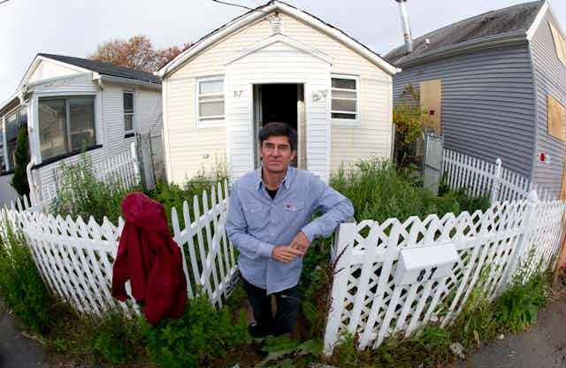 A man stands in front of a small cottage next to a boarded up home. The yard is filled with weeds inside a white picket fence. 