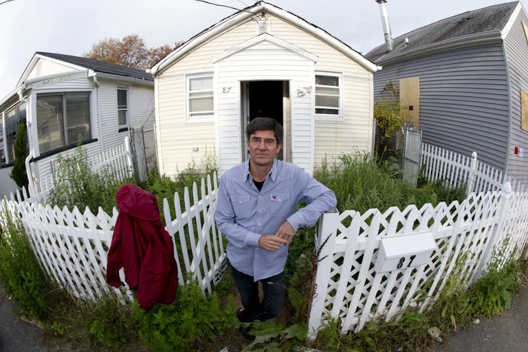 A man stands in front of a small cottage next to a boarded up home. The yard is filled with weeds inside a white picket fence.