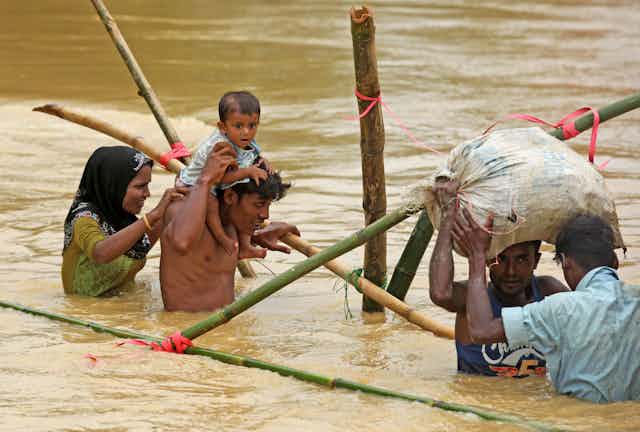 Four Rohingya adults, one carrying an infant on his shoulders, wading through waist high brown water