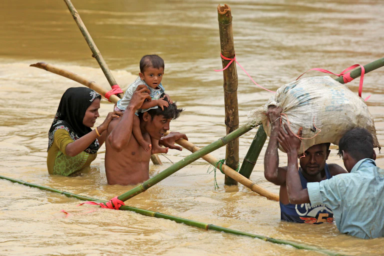 Four Rohingya adults, one carrying an infant on his shoulders, wading through waist high brown water