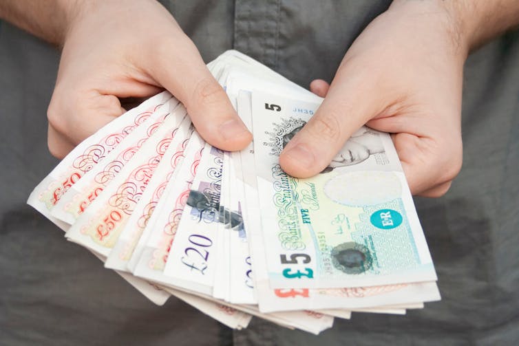 Two hands hold a fan of GBP banknotes: £5, £10, £20, £50.