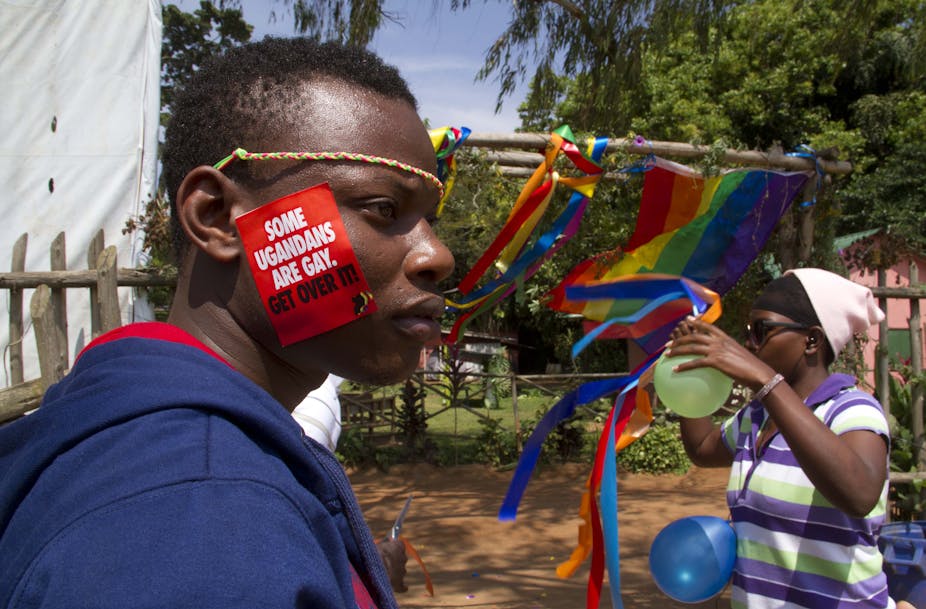 Man with 'Some Ugandans Are Gay. Get Over It' sticker on his face in foreground with man blowing up multicolored balloons in front of rainbow pride flag and rainbow ribbons in background