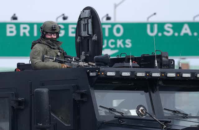 A police officer in green military style fatigues carrying an assault rifle stands atop an armoured vehicle
