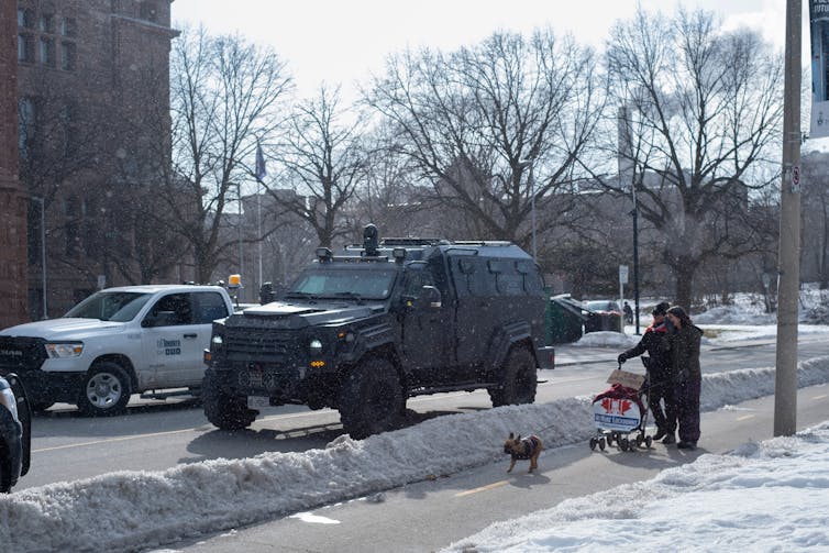 A man with dogs walks passed a black military style armoured vehicle on a snowy city street.
