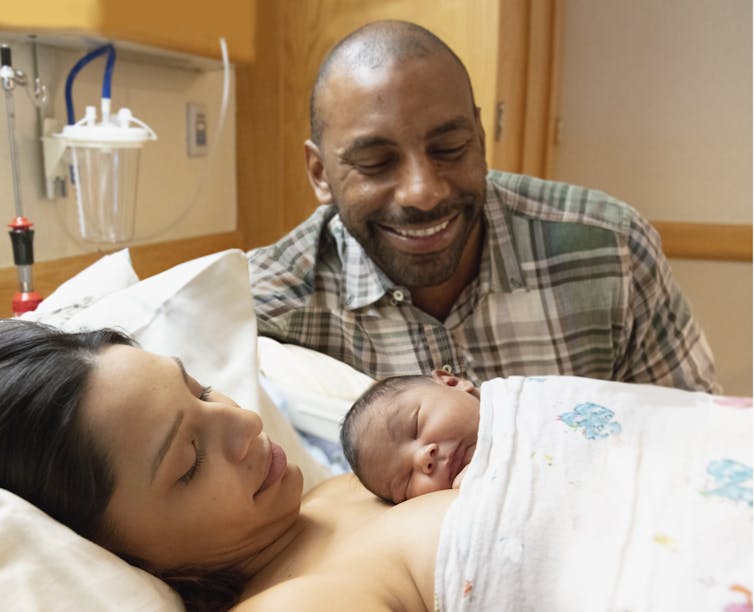 infant on mother's chest in hospital bed with father smiling down at baby