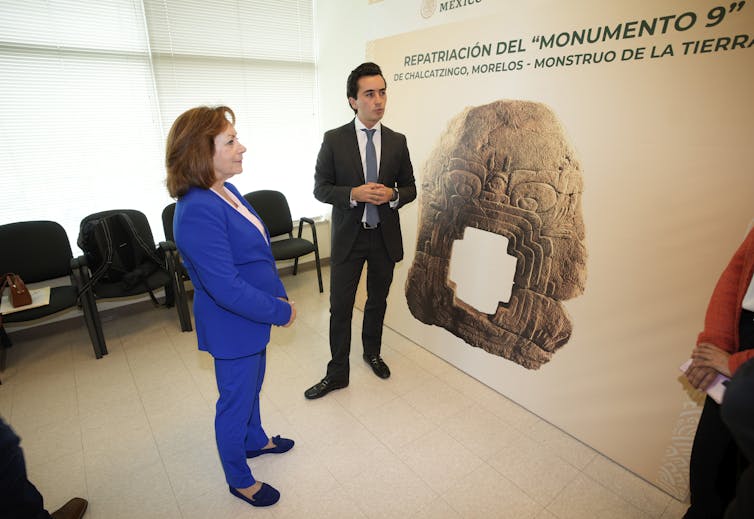 A woman in a bright blue suit and a man in a black suit stand in front of a large poster with a stone statue on it.