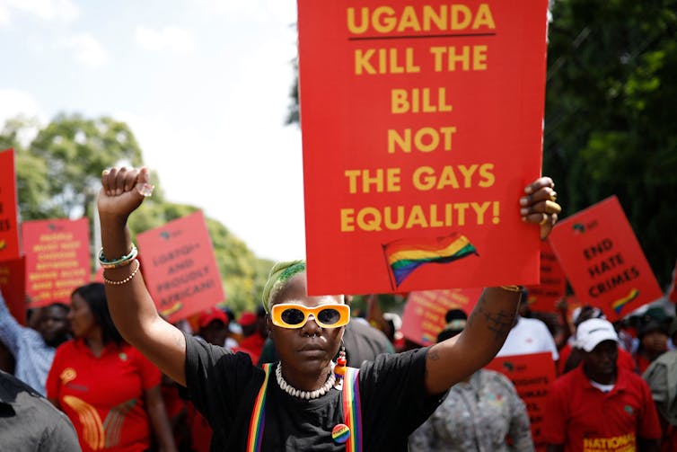 Person wearing yellow sunglasses, taking part in a protest, raises a fist and holds a red and yellow sign saying 'Uganda: Kill the bill not the gays. Equality!'