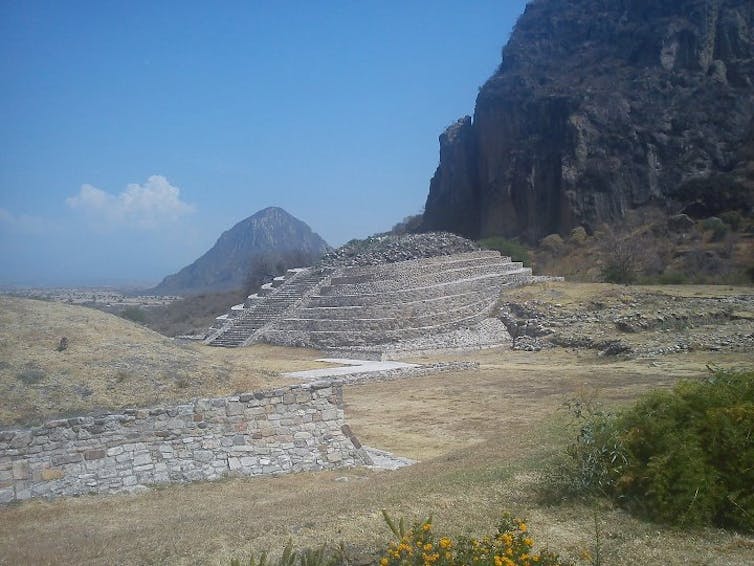 A low stone pyramid-like structure set against a craggy hill and blue sky.