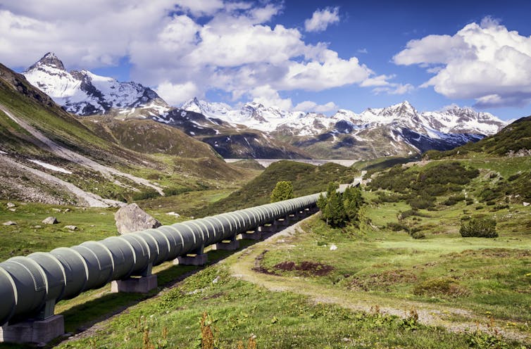 A pipeline runs through a green valley towards mountains and a blue sky with clouds in the background.