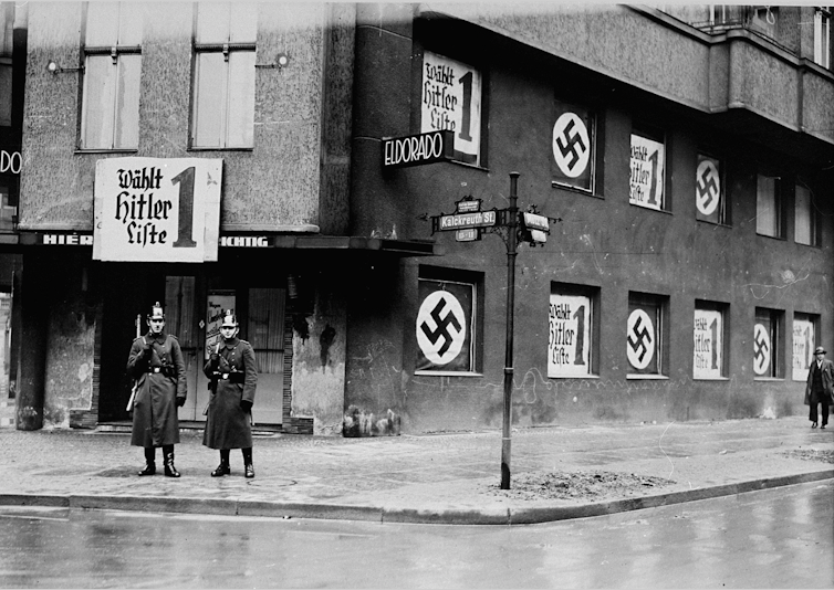 Two police officers stand in front of a shuttered nightclub, which has Nazi banners hanging in the window.