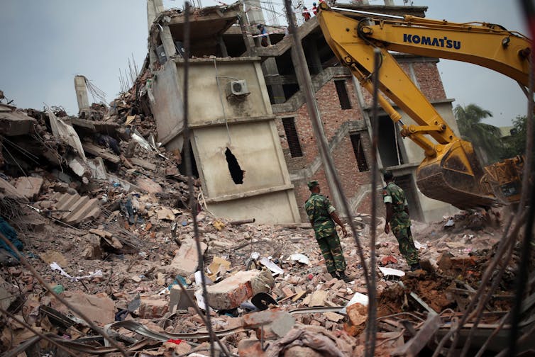 Two men in army fatigues stand on a pile of rubble in front of a collapsed building.