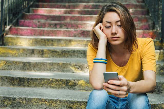 Dressed in a yellow shirt and blue jeans, and sitting on outdoor cement steps, a teenage girl stares at her cellphone.