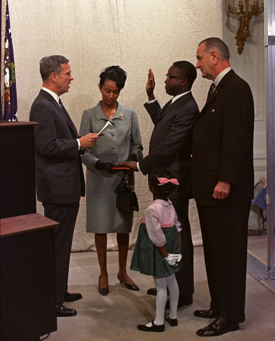 A man with his left hand on a bible held by a woman on his right, holds his right hand face-high, palm side out, as he speaks to a man facing him. Another man and little little girl stand close by on his left.