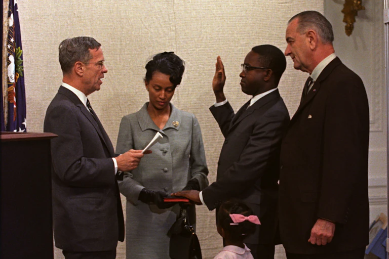 A man with his left hand on a bible held by a woman on his right, holds his right hand face-high, palm side out, as he speaks to a man facing him. Another man and little little girl stand close by on his left.