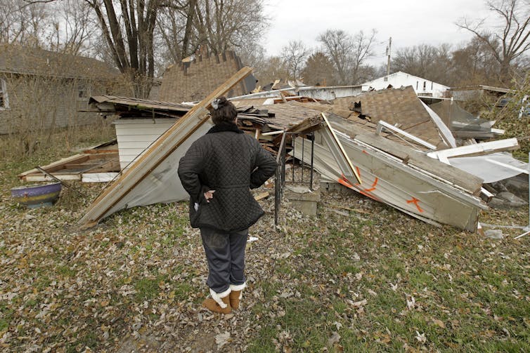 A woman stands in a coat and boots with her hands on her hips, staring at the flattened roof and walls of her former home. It was demolished after the owner agreed to a FEMA buyout. Bright orange X's marked it for demolition.