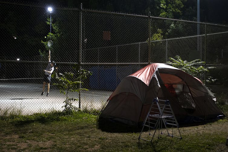 A homeless person's tent sits outside a tennis court where a man plays tennis.