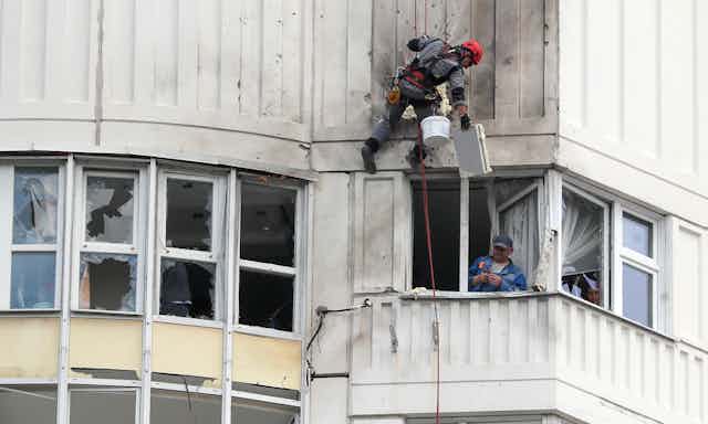 A worker repairs damage to an apartment building in Moscow as the inhabitant looks out.