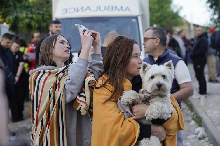 Two women wrapped in blankets stood in front of an ambulance, one holding a dog, the other taking phone footage.