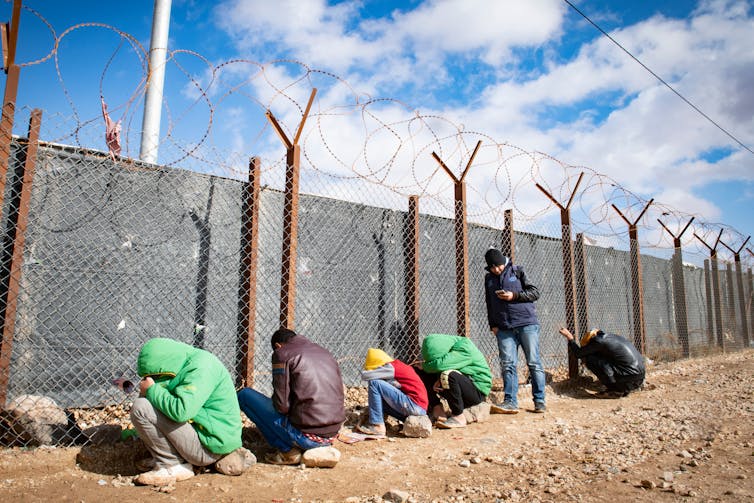 Young men sitting on rocks, wearing jackets and all curled into themselves, next to a tall barbed wire fence