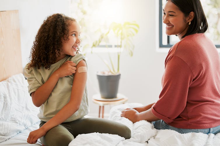 A girl proudly shows her mother a plaster on her upper arm.