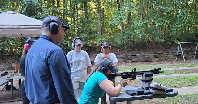 Student shoulders a rifle while professor and fellow students look on.