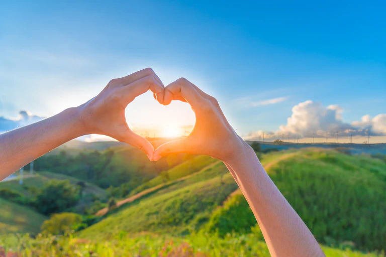 Manos en forma de corazón sobre el sol en un paisaje verde.