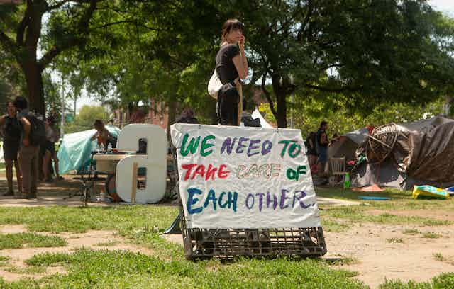 A woman stands next to a sign that reads: we need to take care of each other.