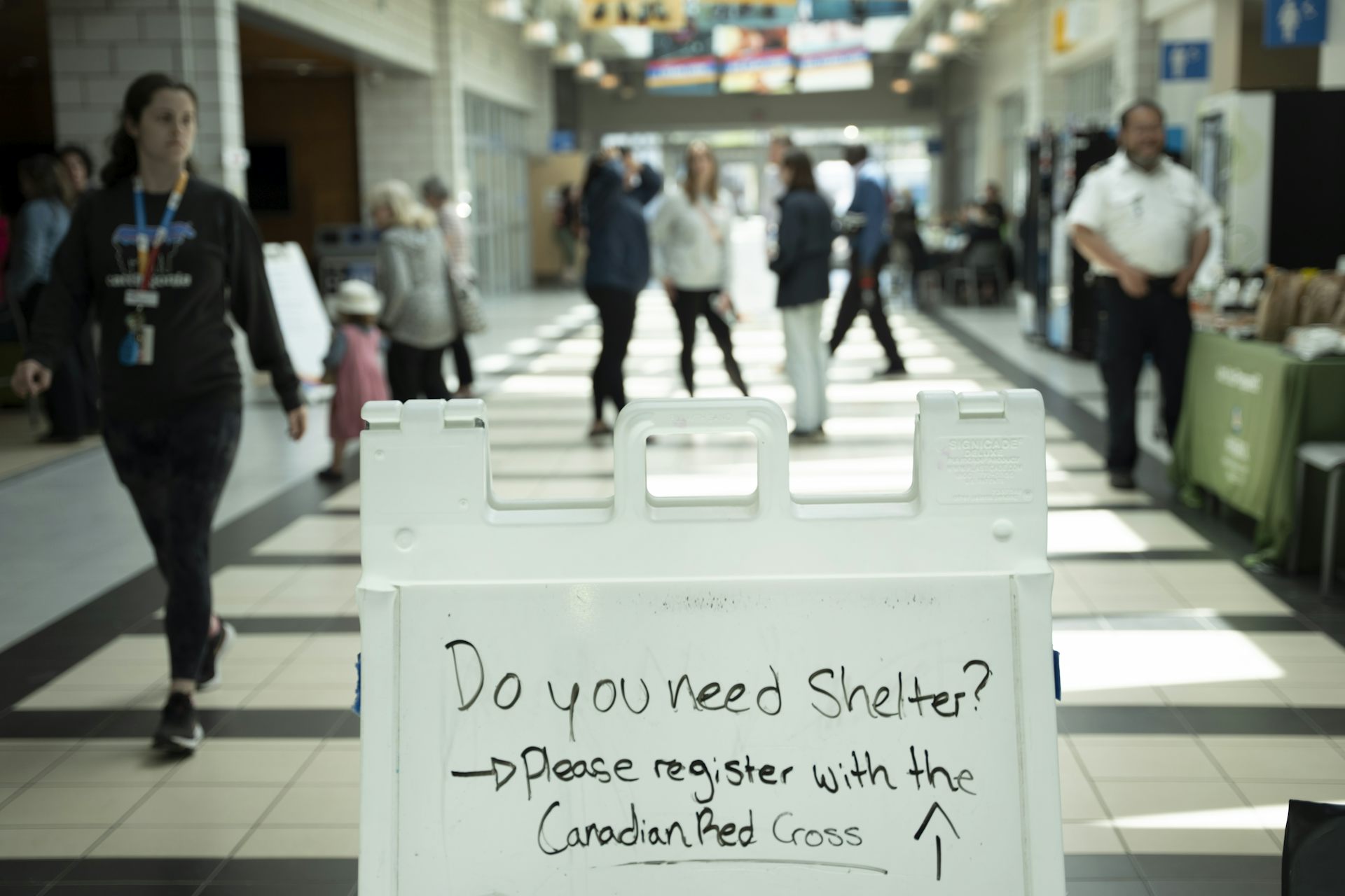 People in a wide corridor, with a sign in the foreground reading 'Do you need shelter? Please register with the Canadian Red Cross.'