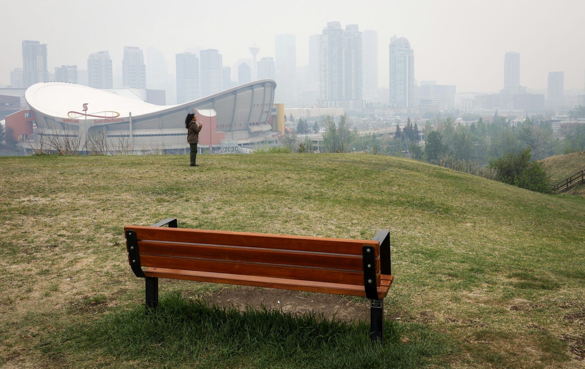 A bench in the foreground with the Calgary Saddledome and downtown blanketed in smoke in the background.