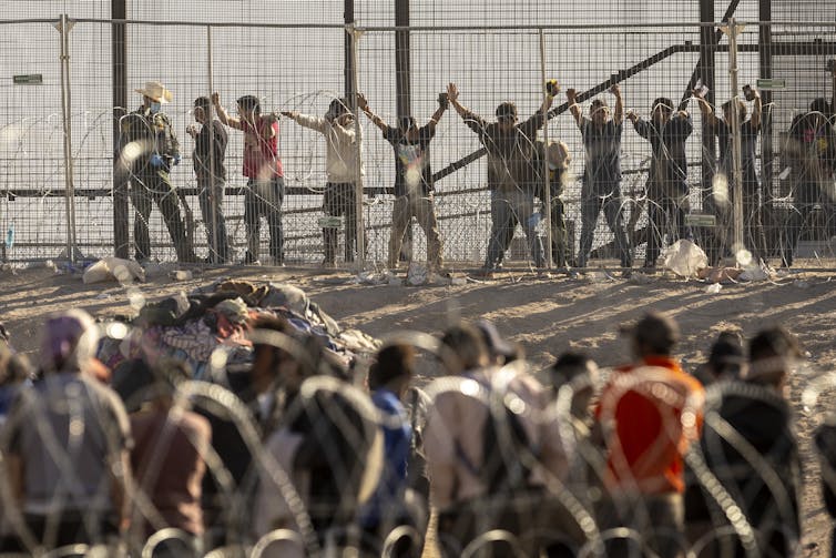 People are seen lined up with their hands up high on a large fence, with rows of other people waiting near the fence.