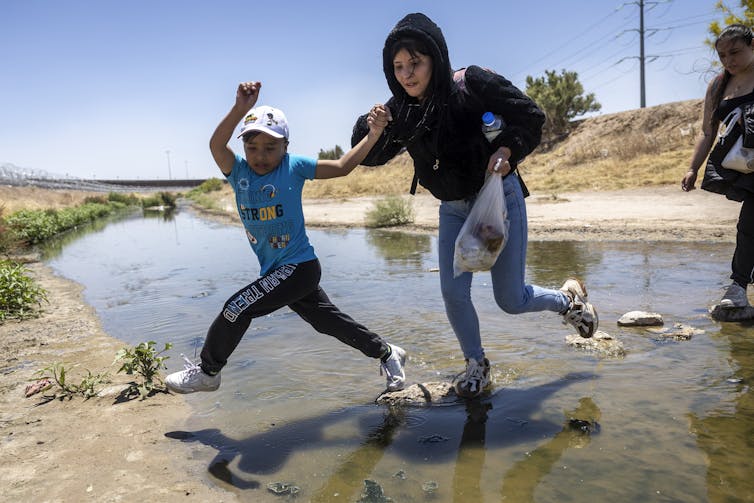 A boy and a woman holding his hand jump across a small river.