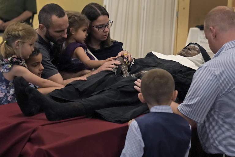 Several people, including children, gather around the body of a nun draped in black clothing.