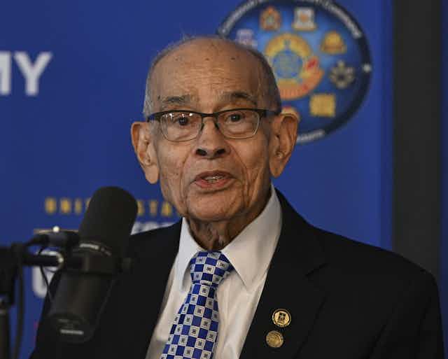An elderly Black man is dressed in a business suit and is speaking near a microphone.