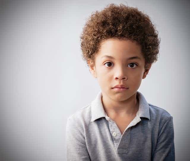 Against a gray background, a sad-looking young boy with curly hair looks at the camera.