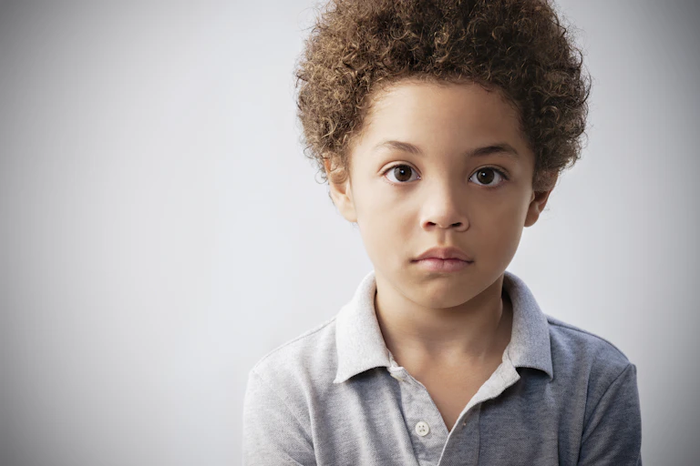 Against a gray background, a sad-looking young boy with curly hair looks at the camera.
