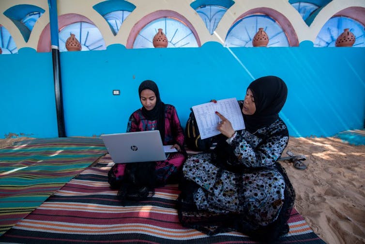 Two women with their heads covered and colorful robes sit on a blanket, holding a laptop and an open notebook.