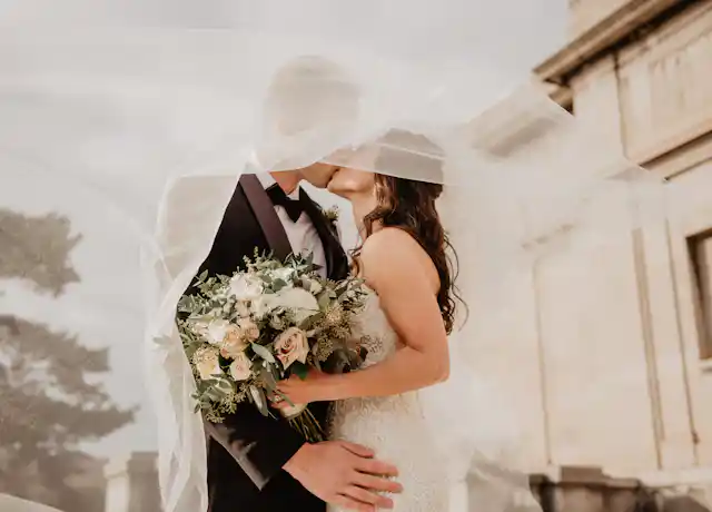 A bride and groom outdoors, kissing under a veil