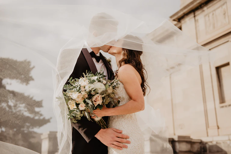A bride and groom outdoors, kissing under a veil