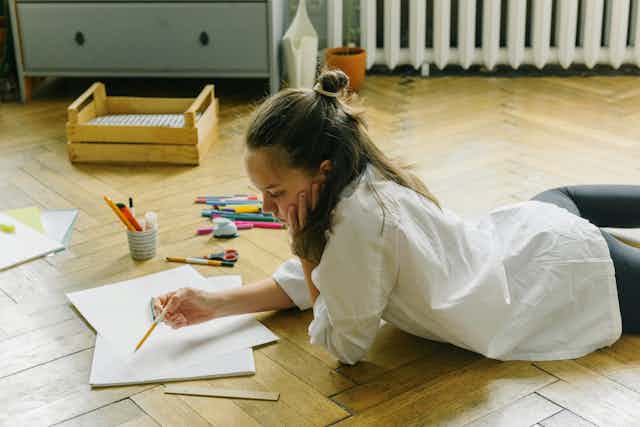 A teenager lies on the floor with paper and pencils. 