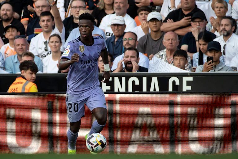 A footballer in a purple strip with the ball in front of fans.