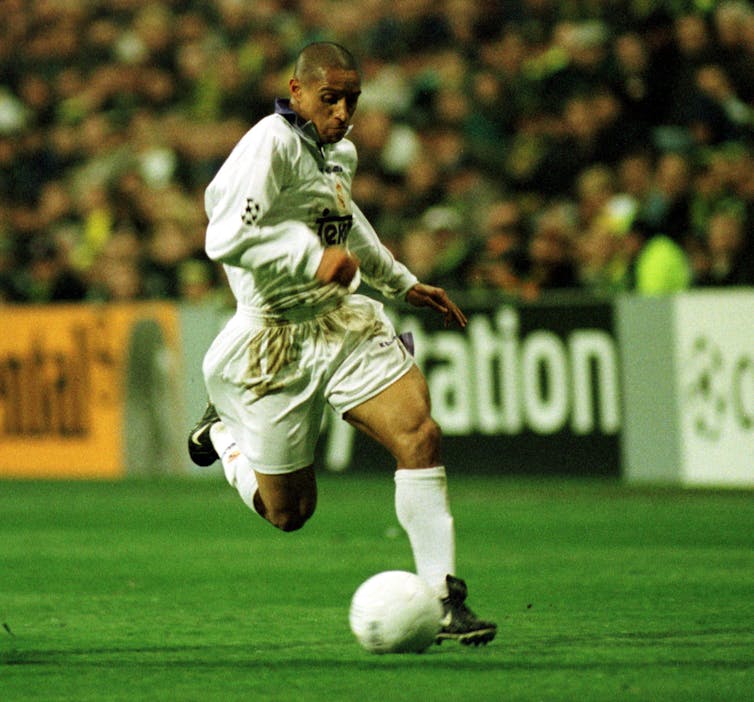 A footballer mid-strike in a white kit on a green pitch.