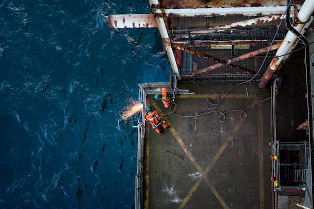 Two workers in orange overalls use cutting torches on the railing of a drilling platform.