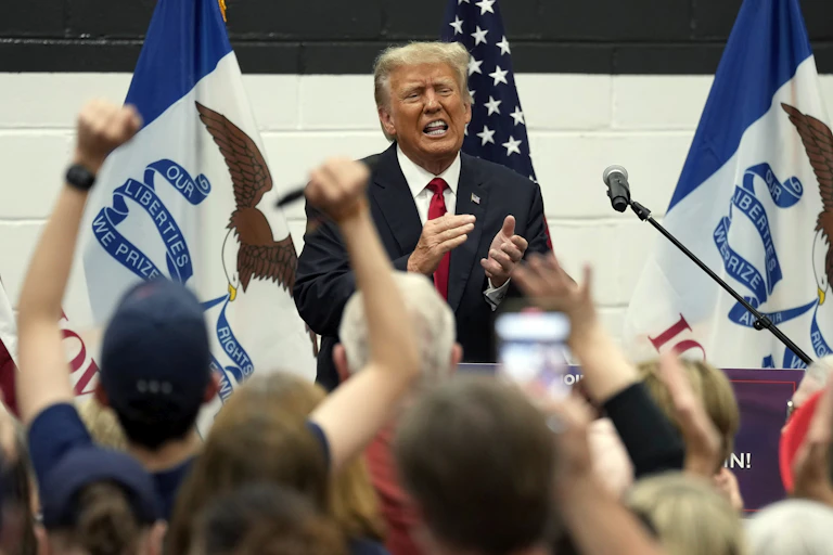 A blonde man in a blue suit and red tie grimaces while clapping as people in front of him raise their arms in support.