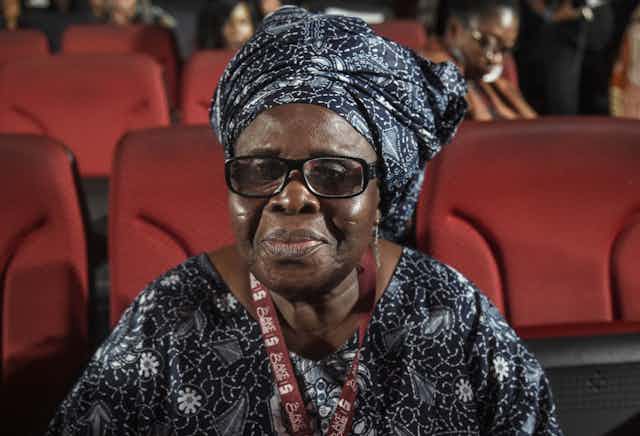 An elderly woman sits in an auditorium with red chairs. She wears traditional attire with an African print headgear and thick-rimmed spectacles, a small smile on her lips.